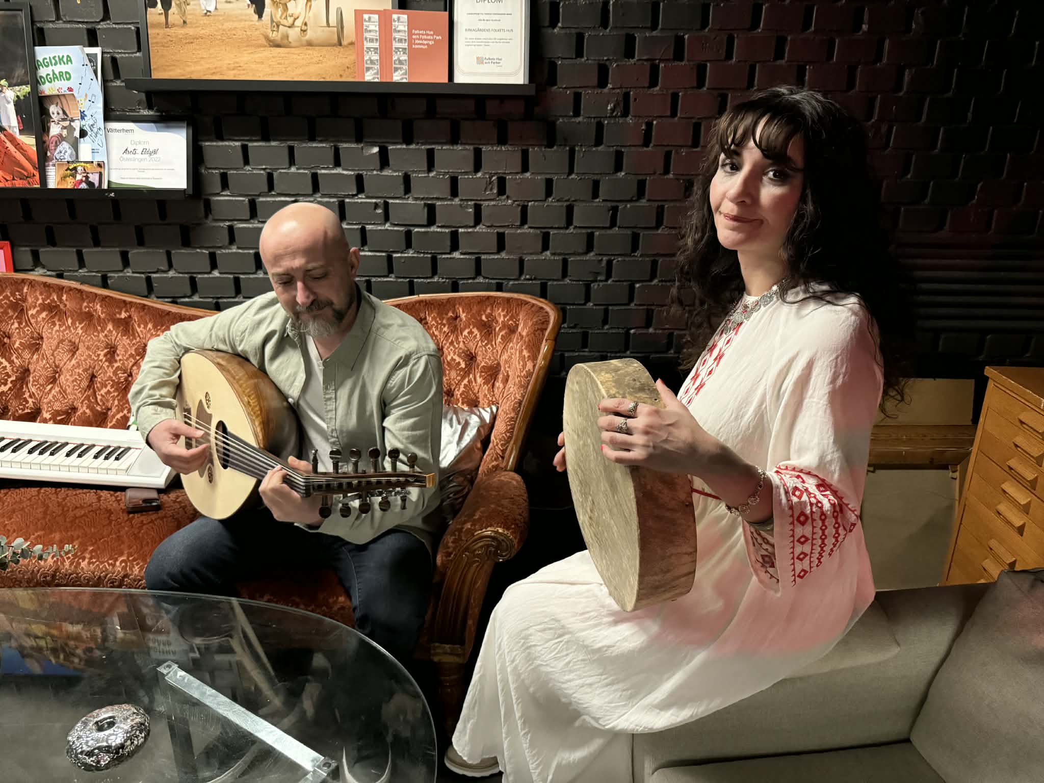 En man spelar oud och en kvinna spelar ramtrumma medan de sitter i en vintagesoffa i ett mysigt rum.