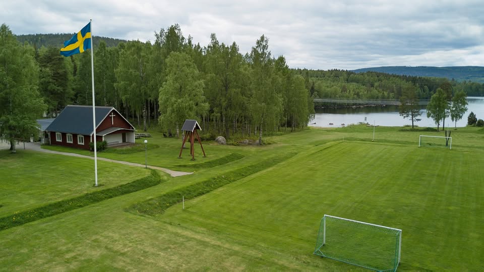 En gräsbevuxen fotbollsplan, ett rött hus och en svensk flagga vid en sjö, omgiven av träd.