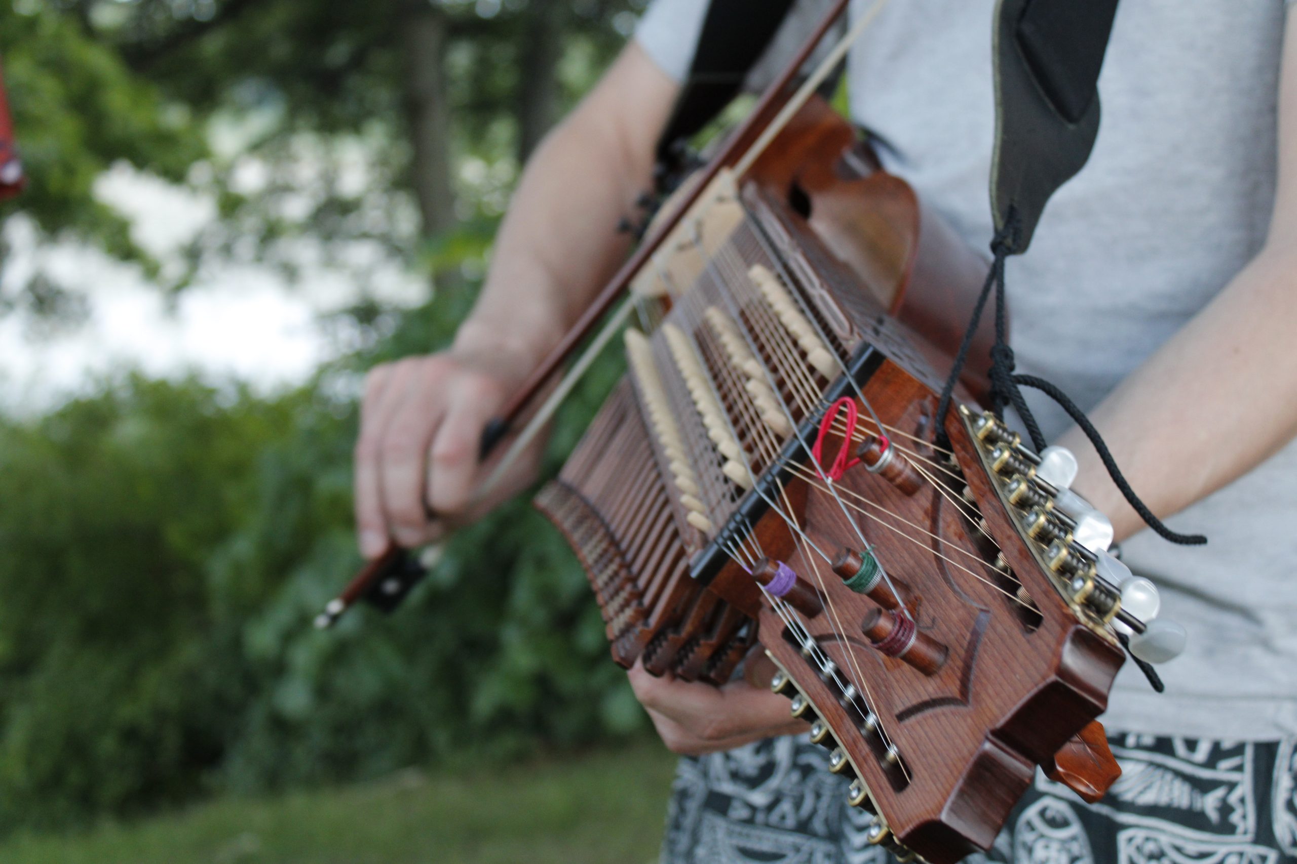 Person spelar nyckelharpa utomhus, med träd och grönska i den suddiga bakgrunden.