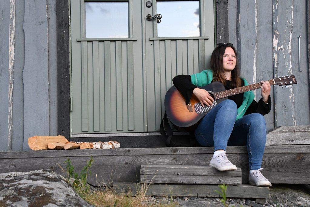 Kvinna sitter på en trätrappa och spelar gitarr, med ved staplad bredvid sig, framför en rustik byggnad.