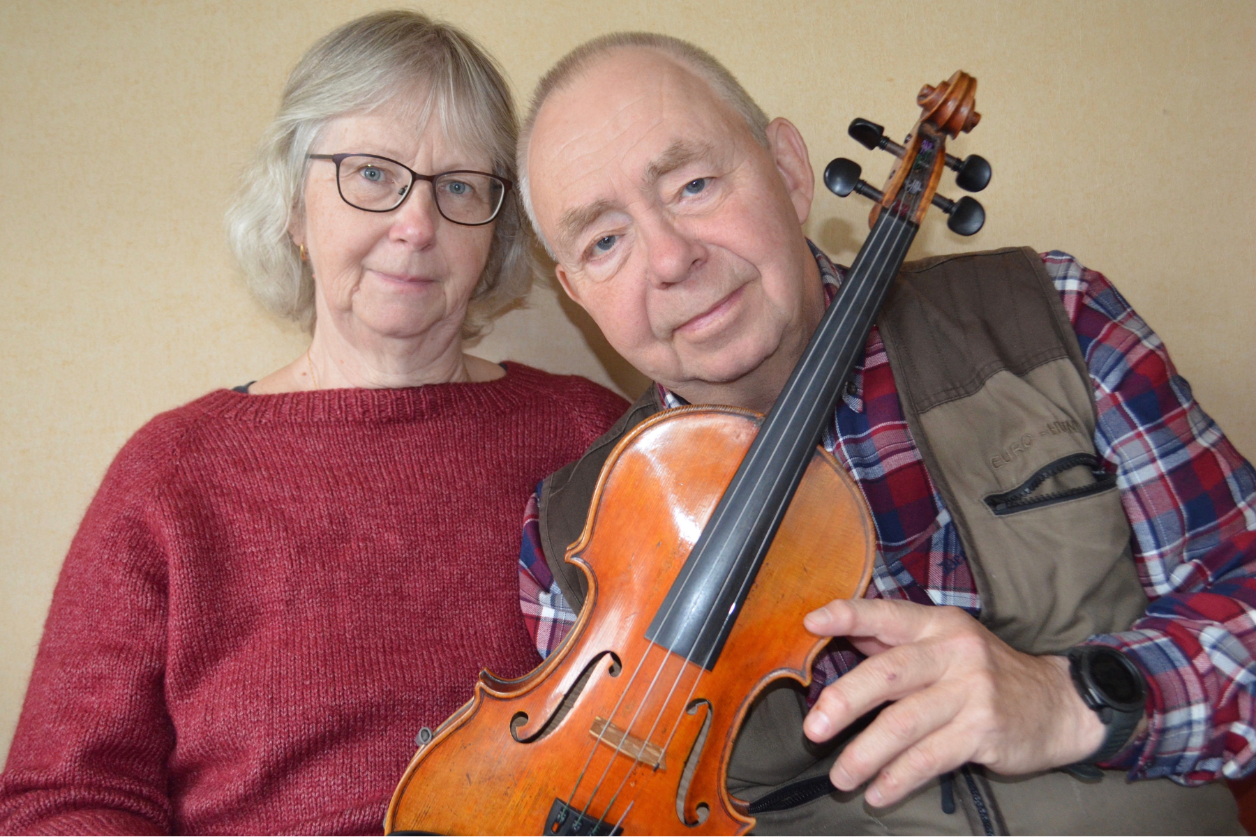 Ett äldre par poserar tillsammans; mannen håller i en violin och ler bredvid kvinnan i röd tröja.