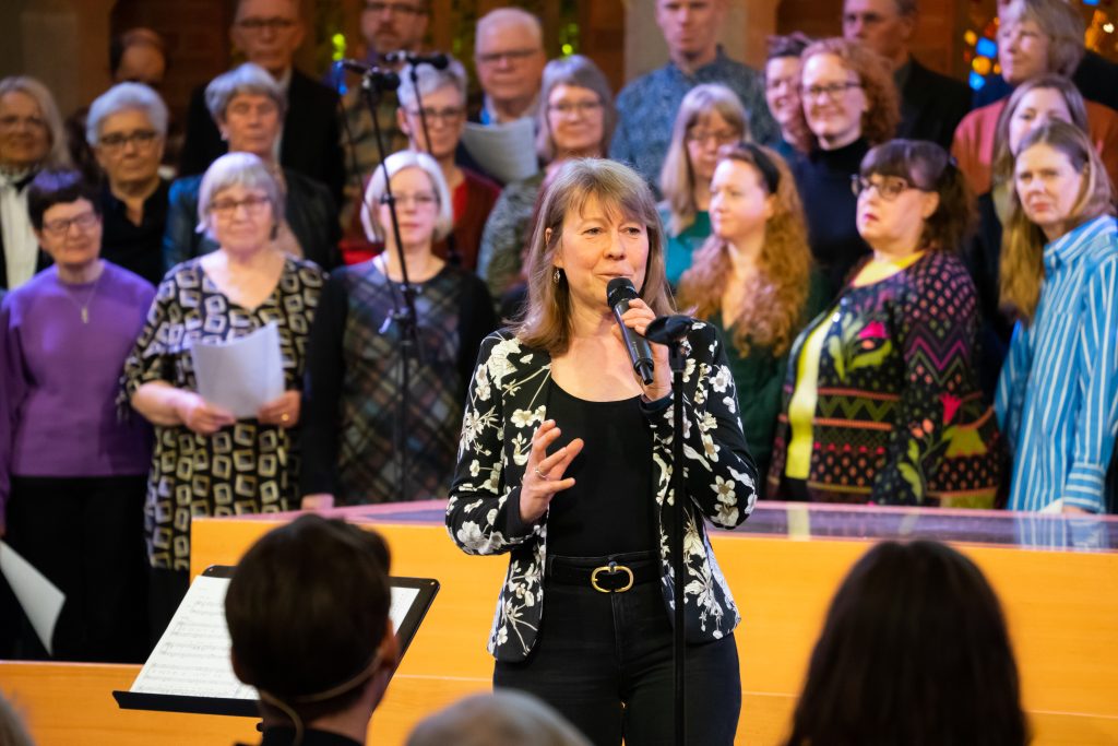 En kvinna sjunger i en mikrofon framför en stående kör i en starkt upplyst inomhusmiljö.