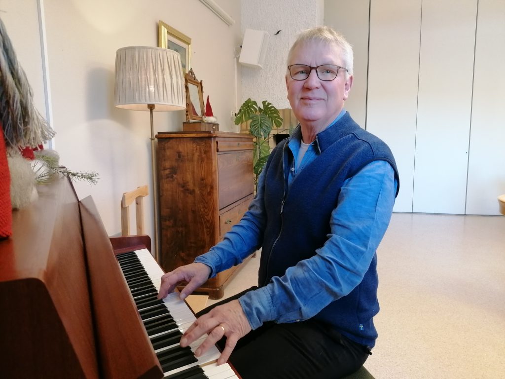 Äldre man i blå skjorta och väst spelar piano i ett mysigt, väl upplyst rum med trämöbler.