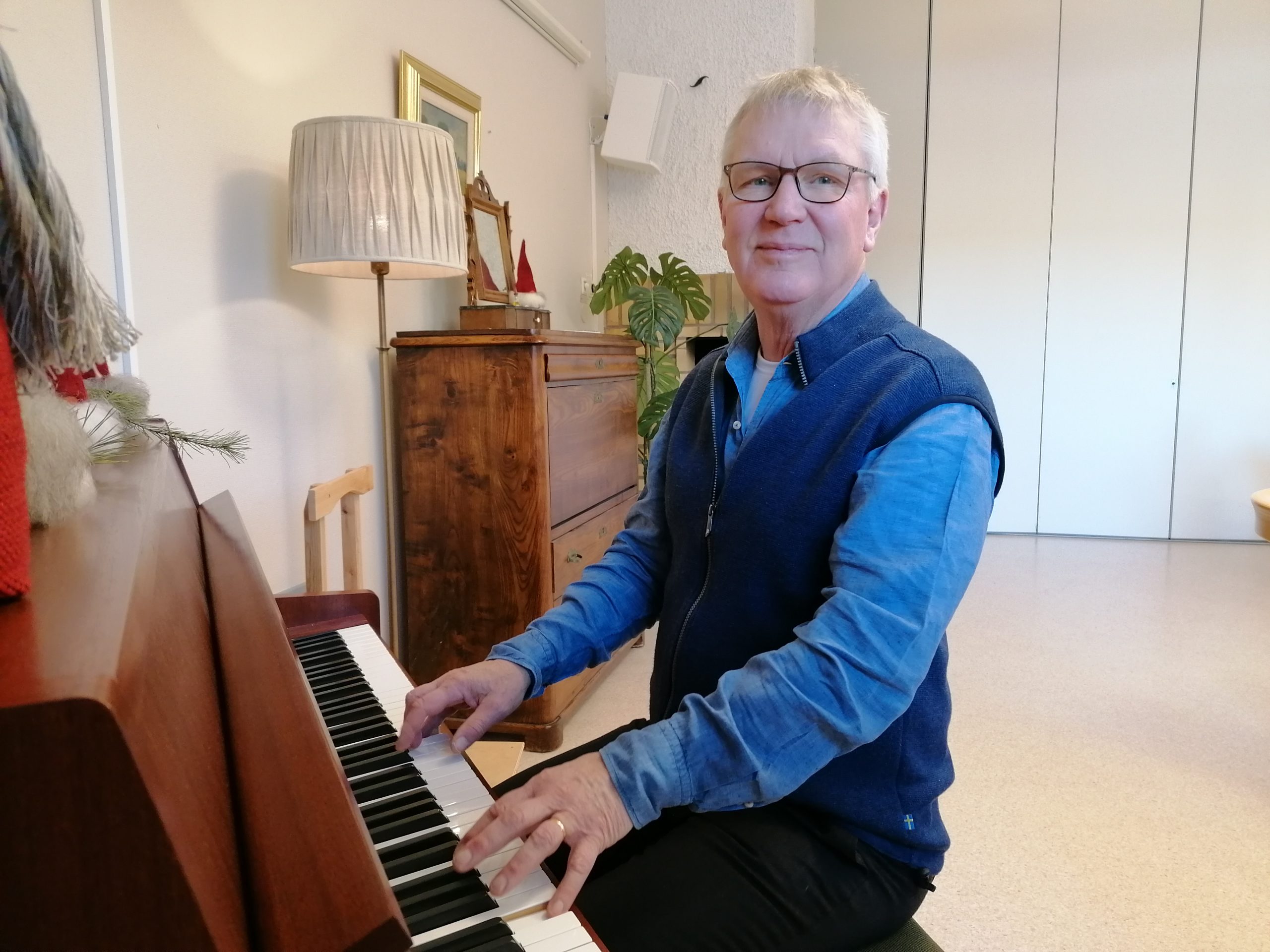 Äldre man i blå skjorta och väst spelar piano i ett mysigt, väl upplyst rum med trämöbler.