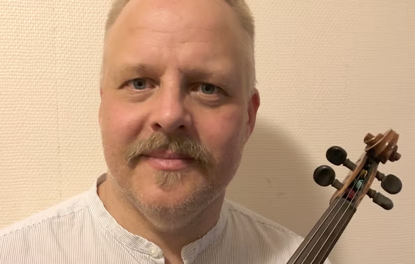 En man med kort blont hår och mustasch håller i halsen på ett stränginstrument och ler mot kameran.