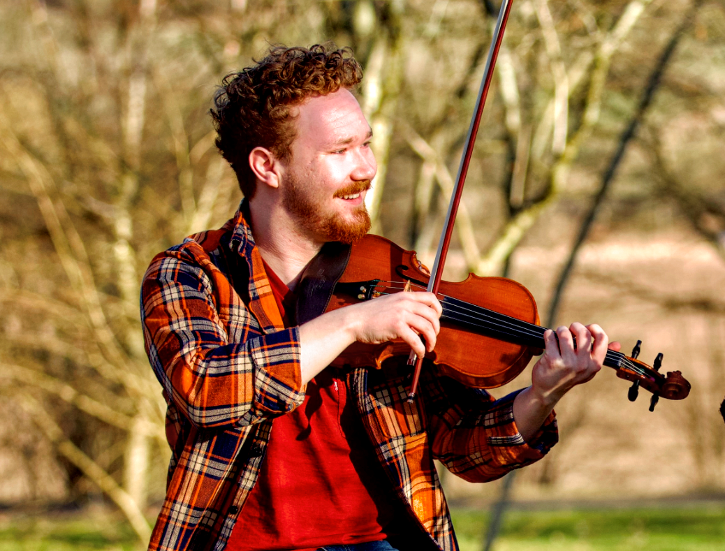En man med lockigt hår spelar violin utomhus och ler i solljuset.