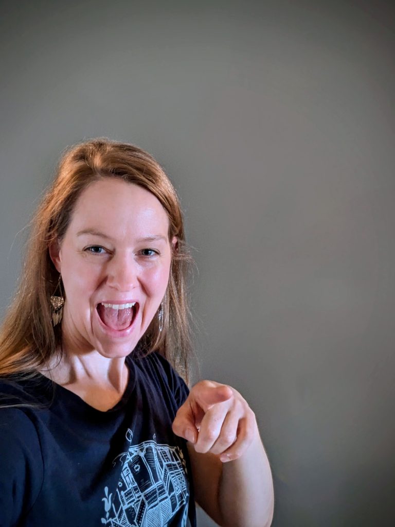 Leende kvinna med långt hår som pekar mot kameran, stående mot en grå bakgrund.