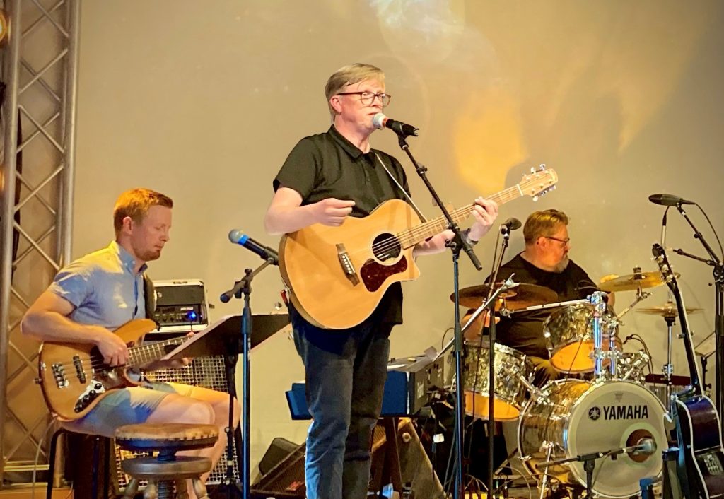 Ett tremannaband spelar på scenen med gitarr, bas och trummor under varm scenbelysning.
