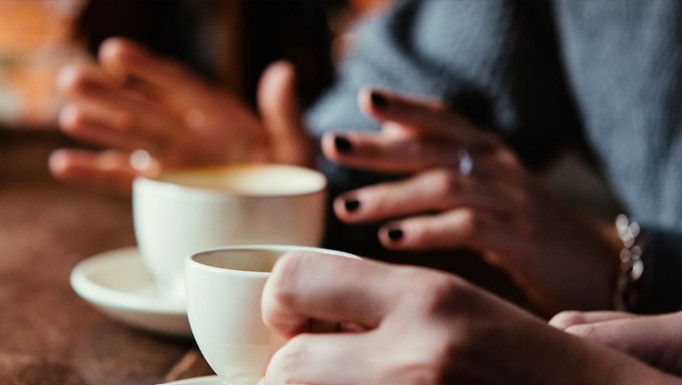 Två personer sitter vid ett bord, håller i vita kaffekoppar och pratar, med fokus på sina händer.