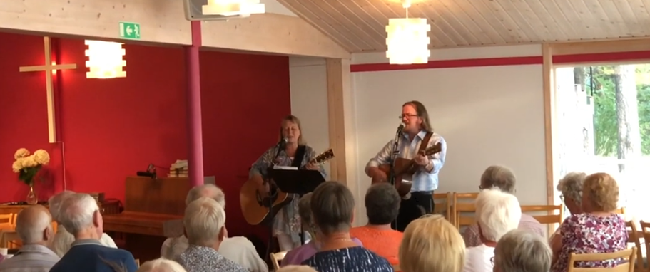 Två musiker spelar gitarr och sjunger för en publik som sitter i ett ljust rum med ett kors på väggen.