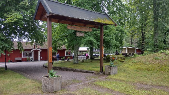 En ingångsbåge i trä med människor och röda byggnader i en grön, trädbevuxen parkmiljö.