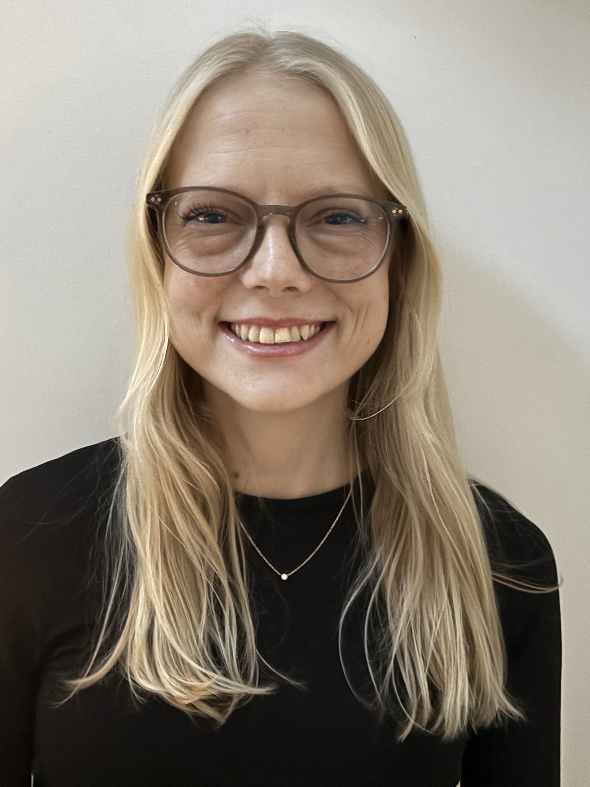 En leende kvinna med långt blont hår, glasögon och en svart topp, stående mot en enfärgad ljus bakgrund.
