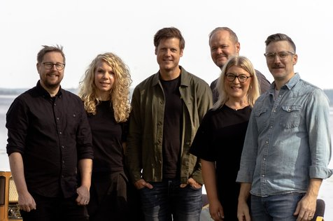Sex vuxna står utomhus i fritidskläder och ler tillsammans mot en ljus, suddig bakgrund.