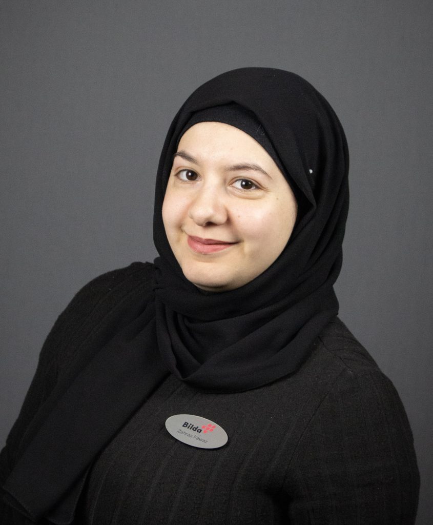 En kvinna i svart hijab och svart klädsel, leende, bär en namnskylt, mot en grå bakgrund.
