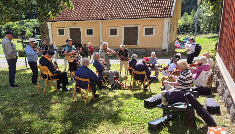 En grupp seniorer som spelar stränginstrument utomhus i en cirkel en solig dag, med folk som tittar på.