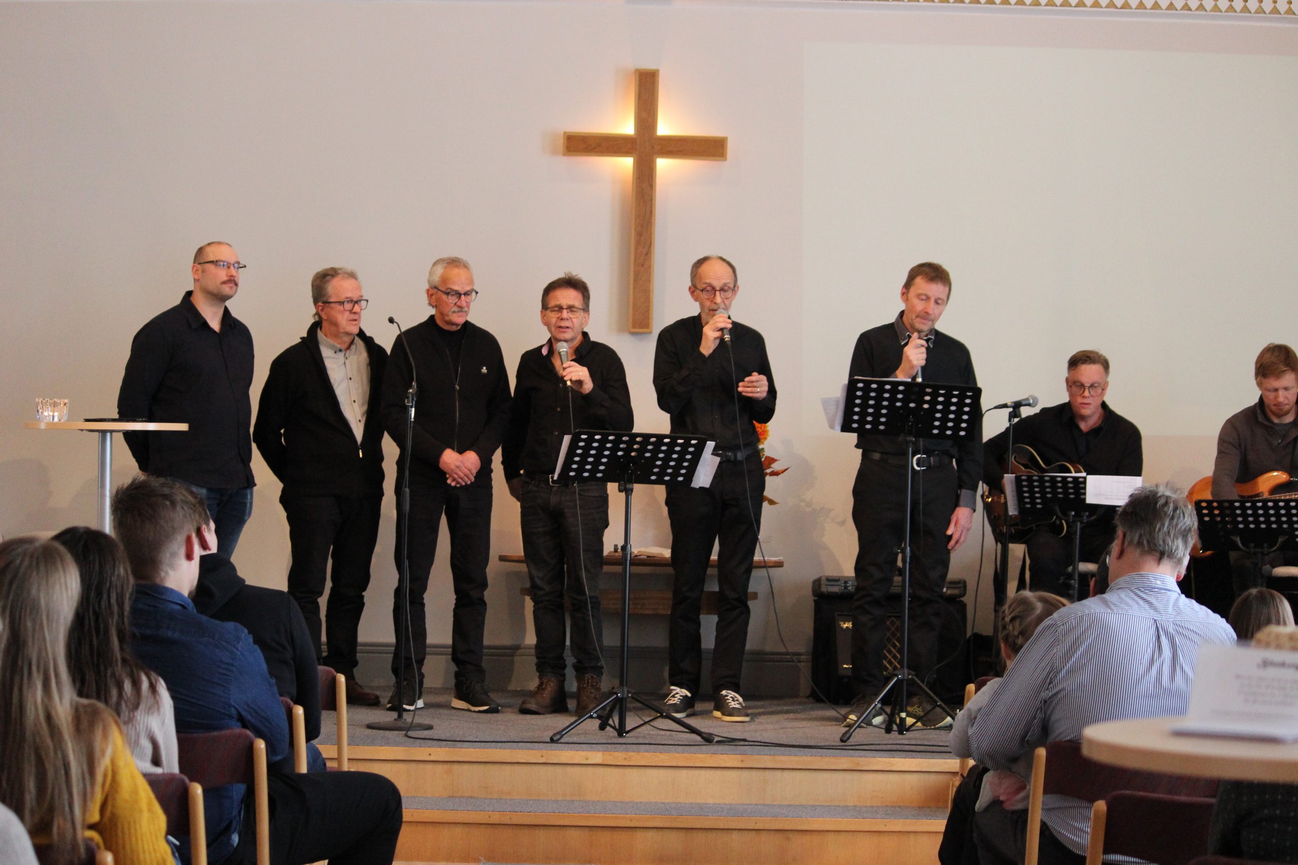 Sju män sjunger på en kyrkoscen med mikrofoner; ett kors är upplyst bakom dem. Publiken är sittande.