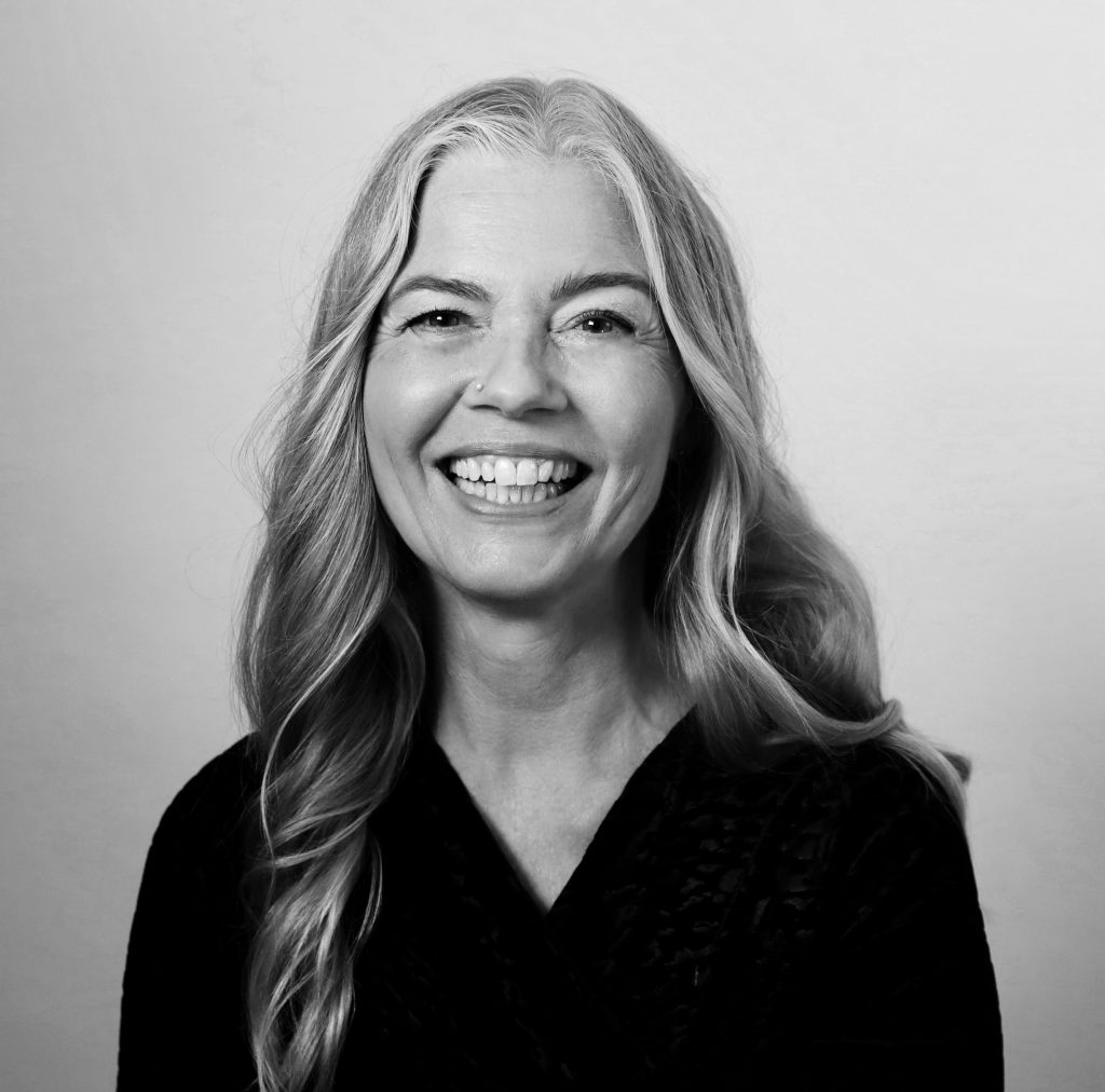 Leende kvinna med långt vågigt hår i svart topp, fotograferad mot en vanlig, ljus bakgrund.