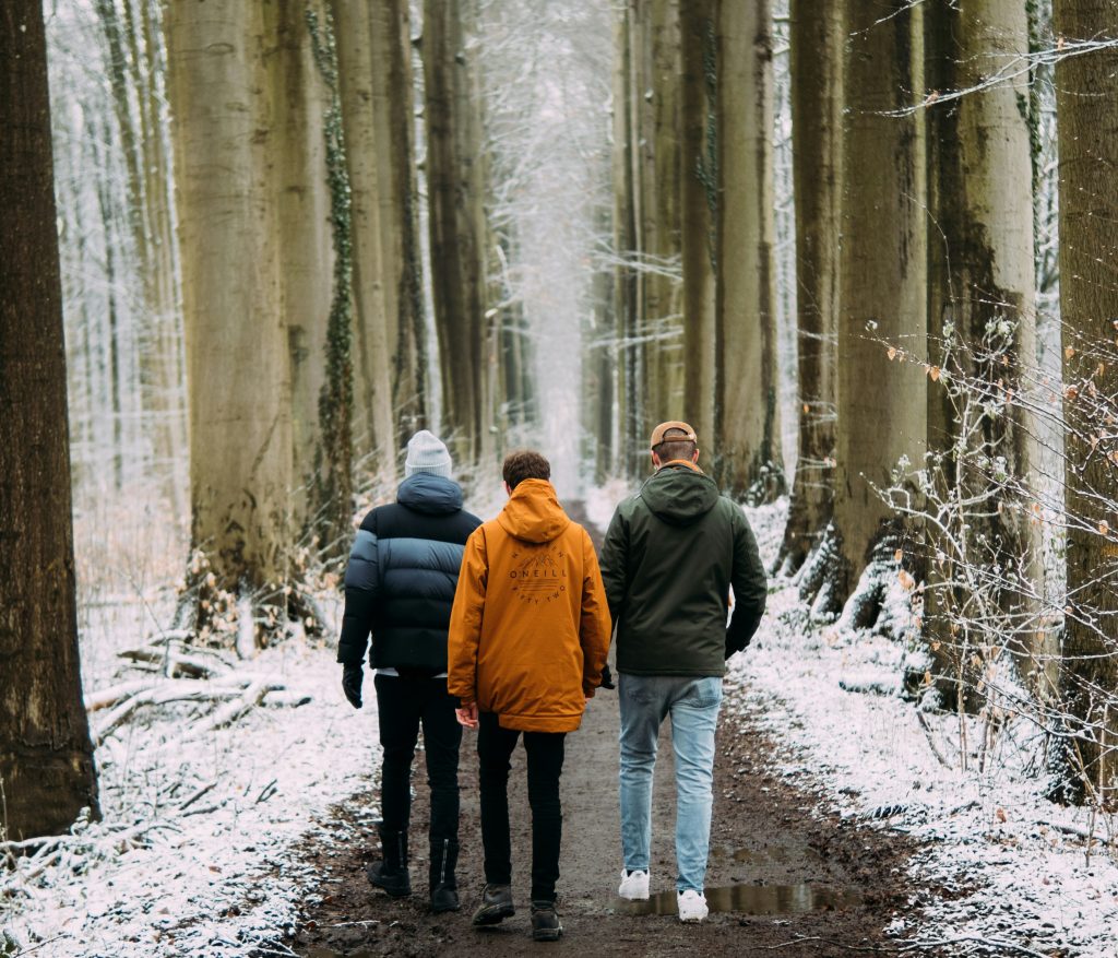 Tre personer går längs en snöig skogsstig, omgiven av höga träd, sett bakifrån.