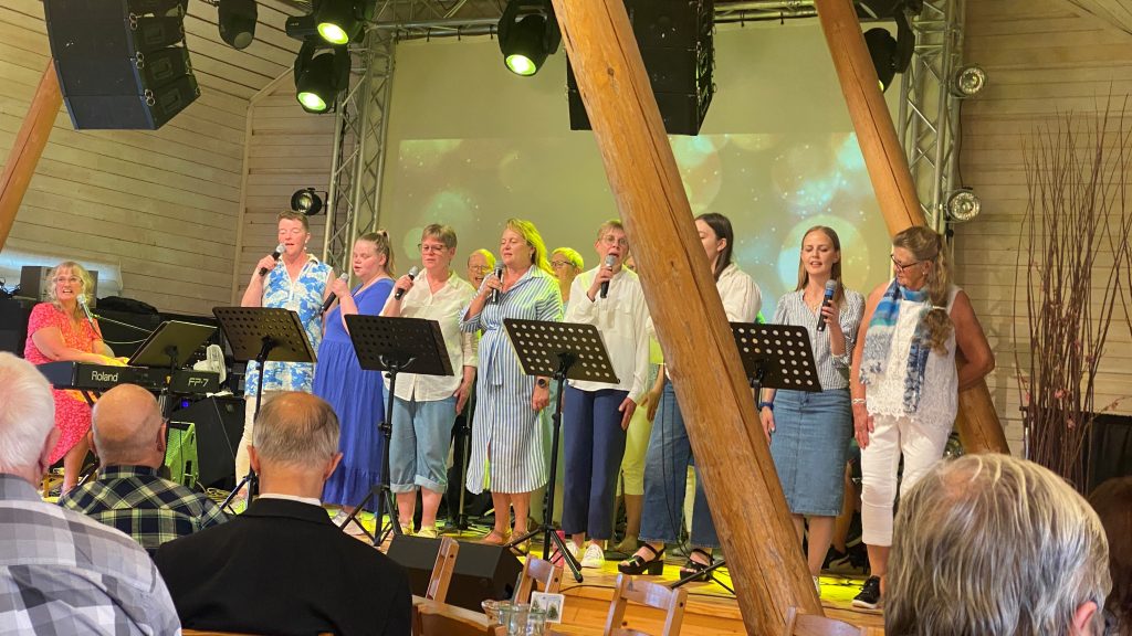 En grupp kvinnor sjunger på scenen med mikrofoner; publiken tittar på från sittande bord i en träpanelförsedd sal.