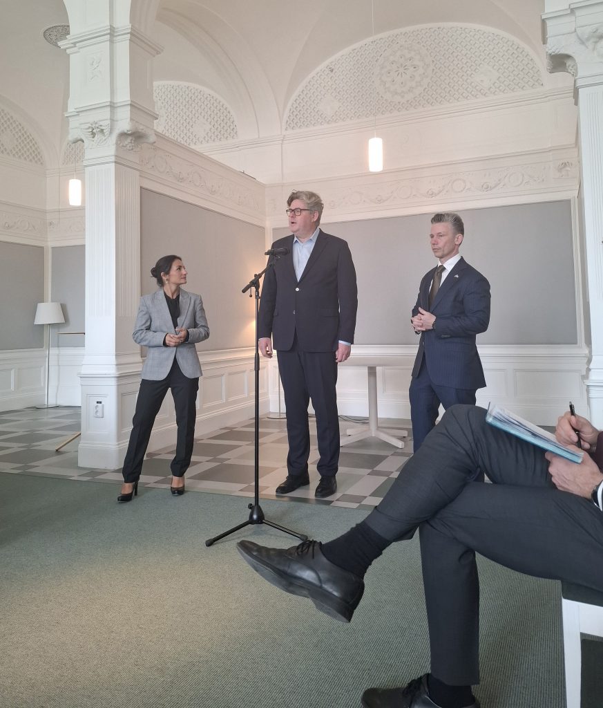 Tre personer från regeringen står och talar i en mikrofon i ett formellt, elegant rum med vita väggar.