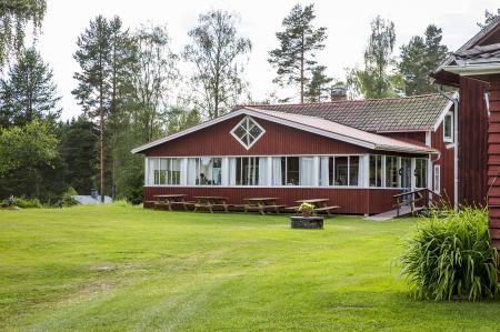 Ett rött trähus med stora fönster, omgivet av gräs, träd och picknickbord utomhus.