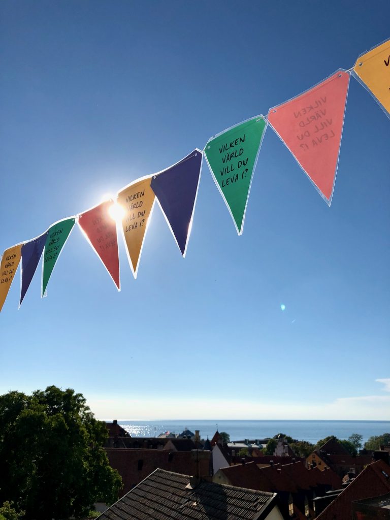 Färgglada trekantiga flaggor med svensk text hänger över hustaken med sol och hav i bakgrunden.