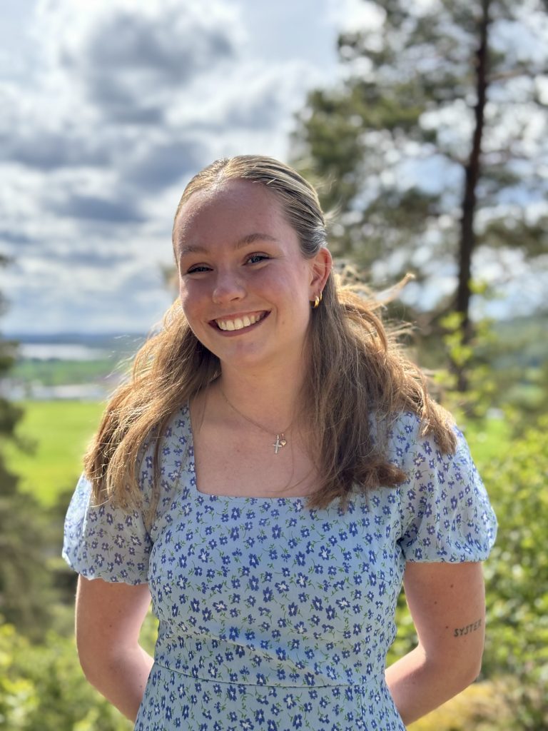 En leende kvinna i en blå blommig klänning som står utomhus med träd och en molnig himmel i bakgrunden.