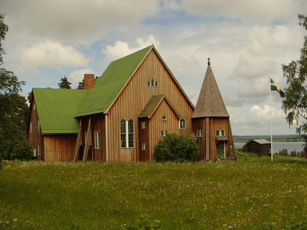 En träkyrka med grönt tak och spir står på en gräsbevuxen äng med en svensk flagga i närheten.