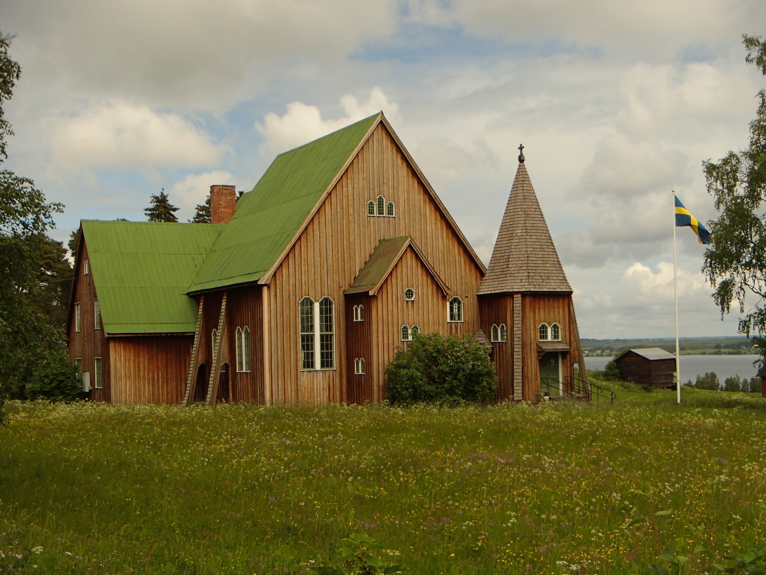 En träkyrka med grönt tak och spir står på en gräsbevuxen äng med en svensk flagga i närheten.