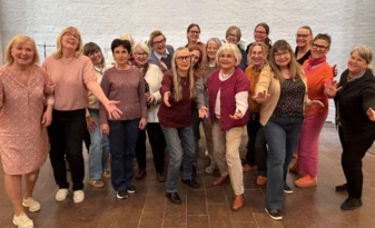 En grupp leende kvinnor poserar tillsammans inomhus, några med utsträckta armar, i en avslappnad och glad miljö.