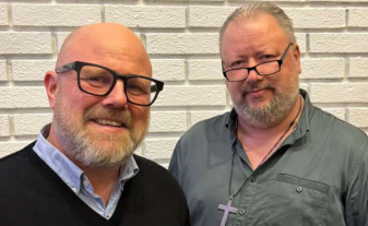 Två leende män med glasögon står framför en vit tegelvägg.