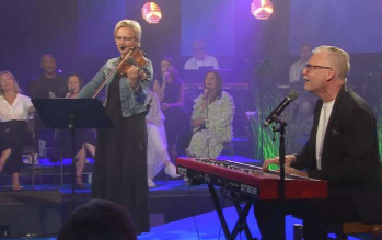 En kvinna spelar violin medan en man spelar keyboard på scenen; kören sjunger i bakgrunden.