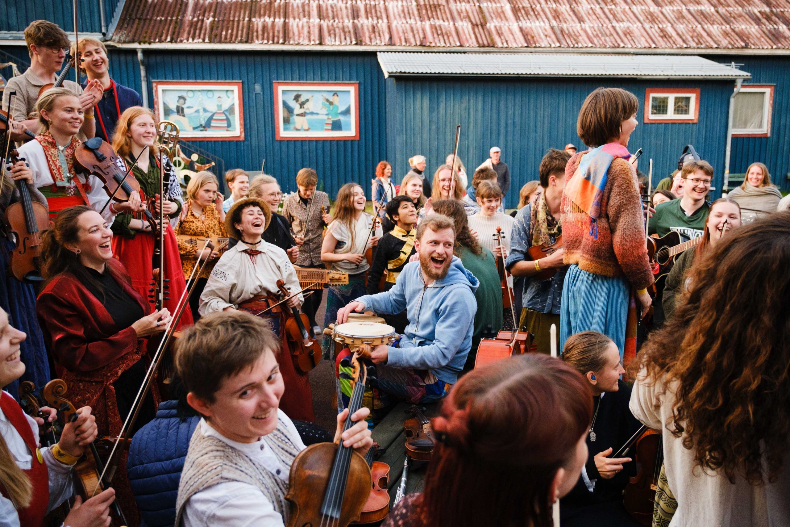 En glad grupp musiker spelar instrument och skrattar tillsammans utomhus i närheten av en blå byggnad.