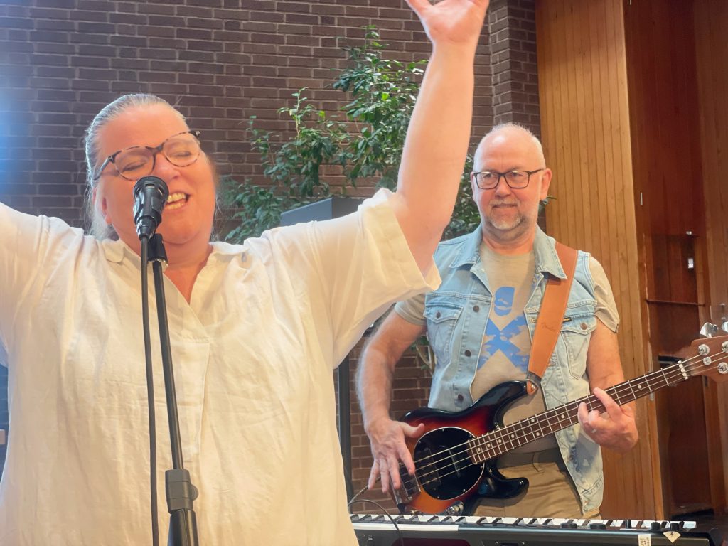 En kvinna sjunger glatt i en mikrofon medan en man spelar basgitarr bredvid henne inomhus.