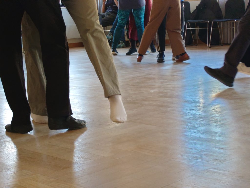 Människor som dansar inomhus, där ben och fötter syns i rörelse på ett polerat trägolv.