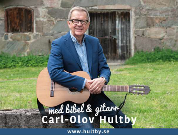 En leende man i blå kostym sitter utomhus med en akustisk gitarr, med en stenmur bakom sig. Text: Carl-Olov Hultby.