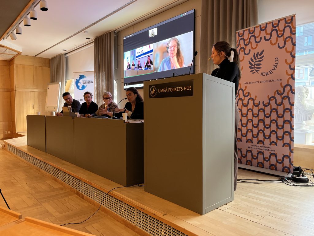 En panel bestående av fem personer sitter vid ett bord, där en person talar från ett talarstativ och ett videosamtal visas på skärmen.