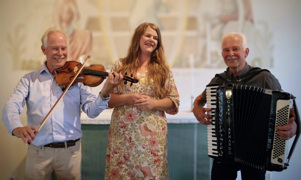 Tre leende vuxna spelar musik tillsammans; en med fiol, en med dragspel och en som sjunger.