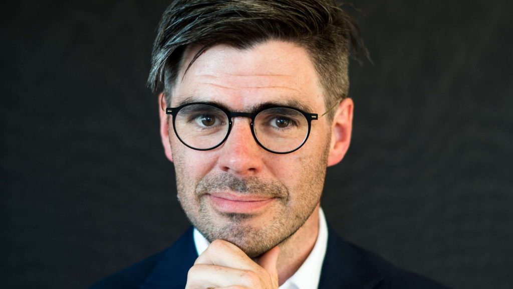 En man med glasögon och mörkt hår, klädd i kostym, som tittar in i kameran med handen mot hakan mot en mörk bakgrund.