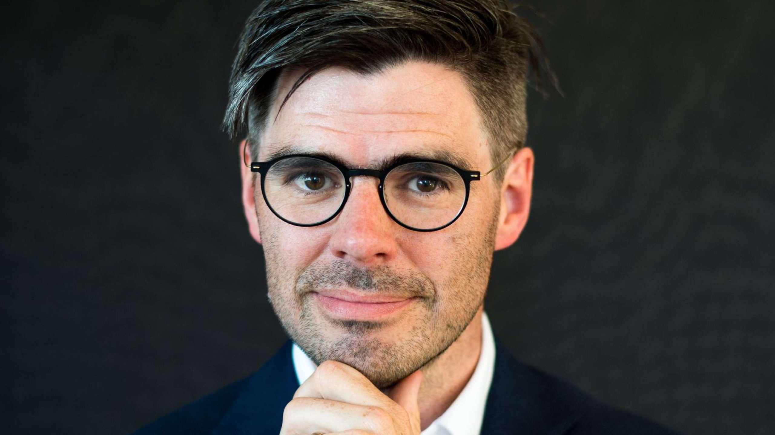 En man med glasögon och mörkt hår, klädd i kostym, som tittar in i kameran med handen mot hakan mot en mörk bakgrund.