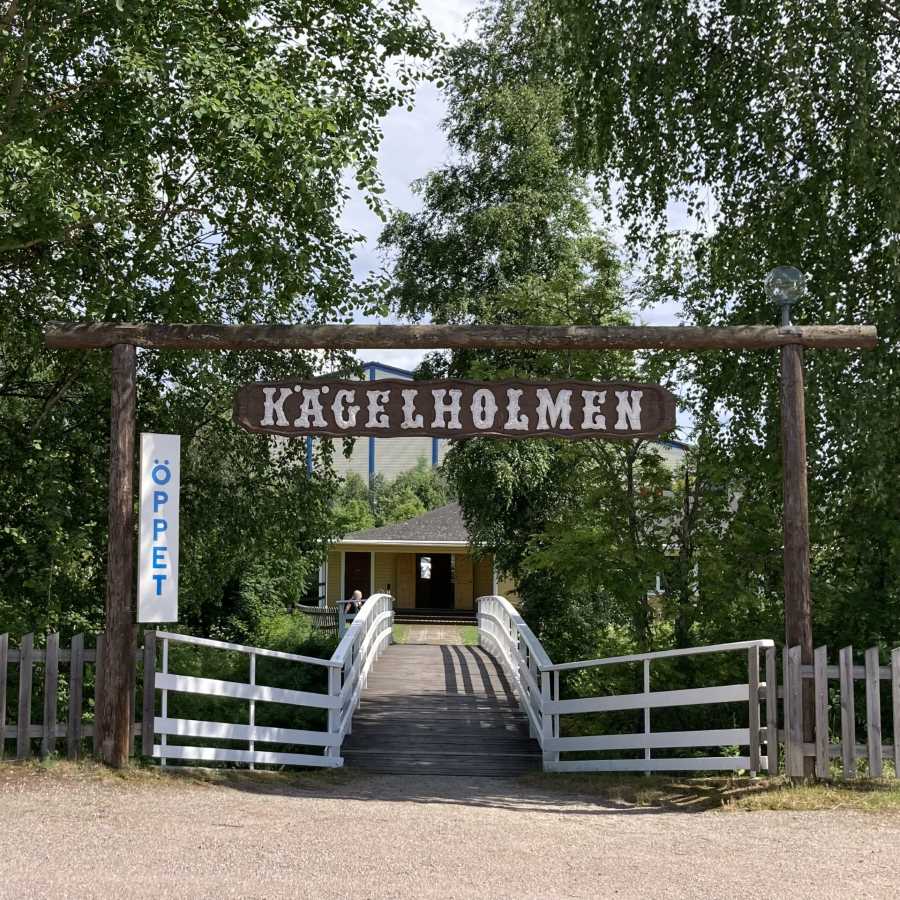 Träskylt med texten "Kagelholmen" över en port med en bro, träd och en gul byggnad i bakgrunden.
