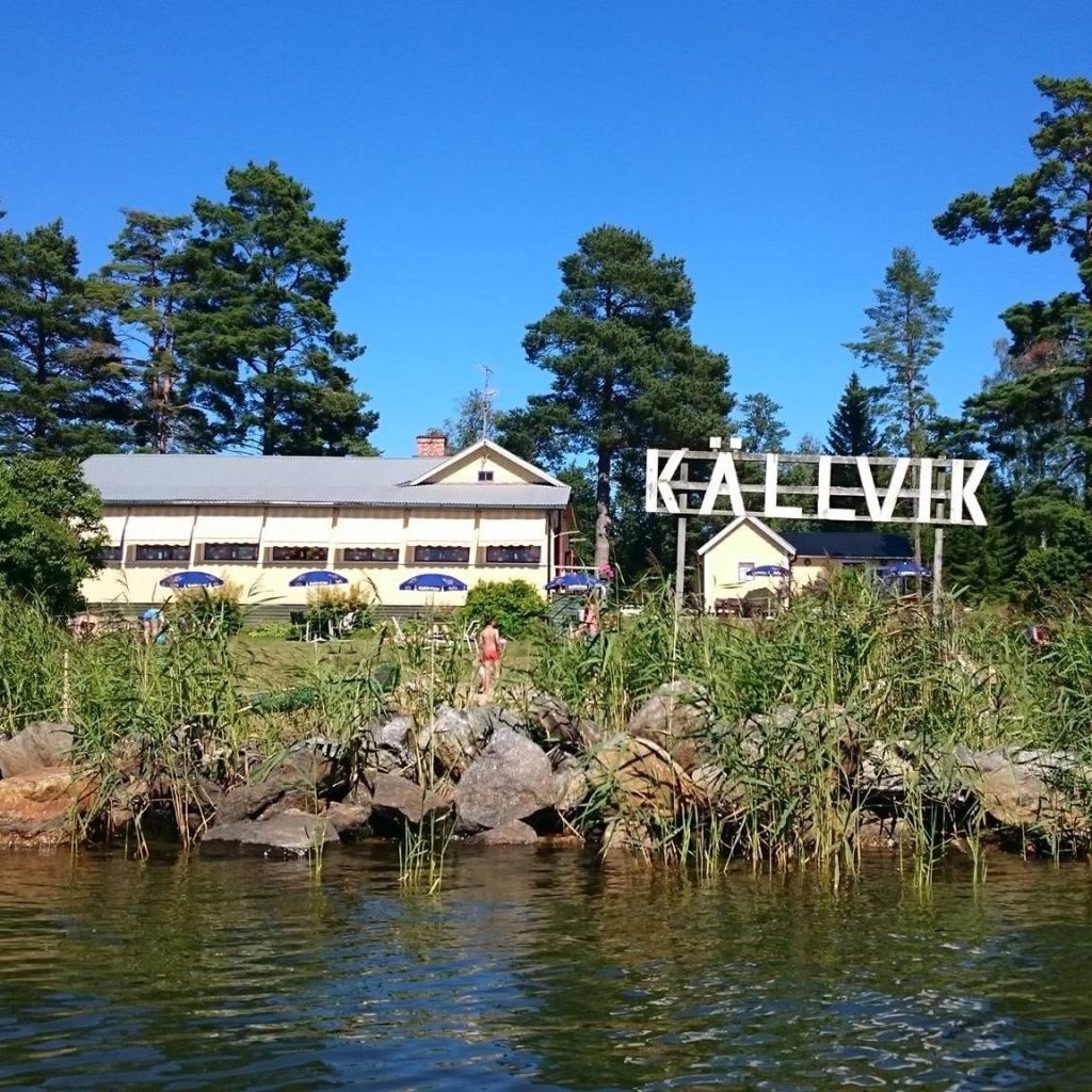 En byggnad vid en sjö med träd, stenar och en stor skylt med texten "KÄLLVIK" under en klarblå himmel.