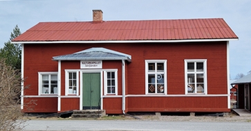 En röd träbyggnad med en grön dörr, vita lister och en skylt ovanför ingången.