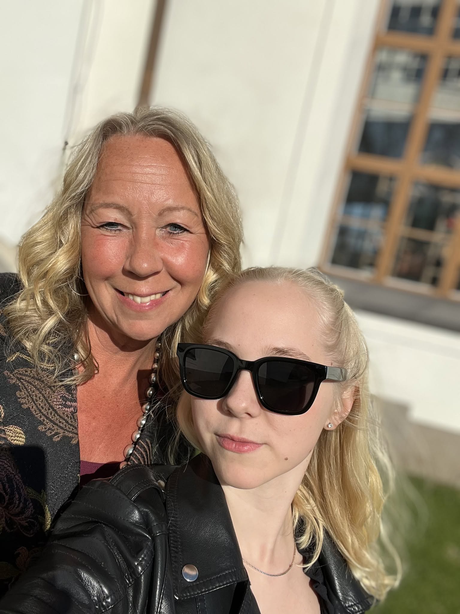 Två kvinnor ler utomhus; den ena bär solglasögon och läderjacka, båda har blont hår och lockiga frisyrer.