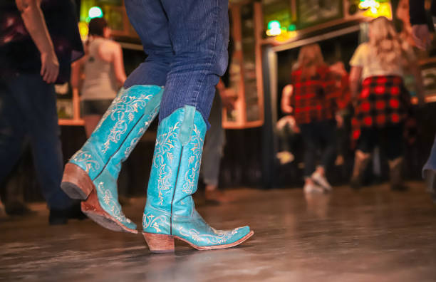 Närbild på blå cowboystövlar medan människor dansar linedance inomhus; i bakgrunden syns dansare i rörelse.