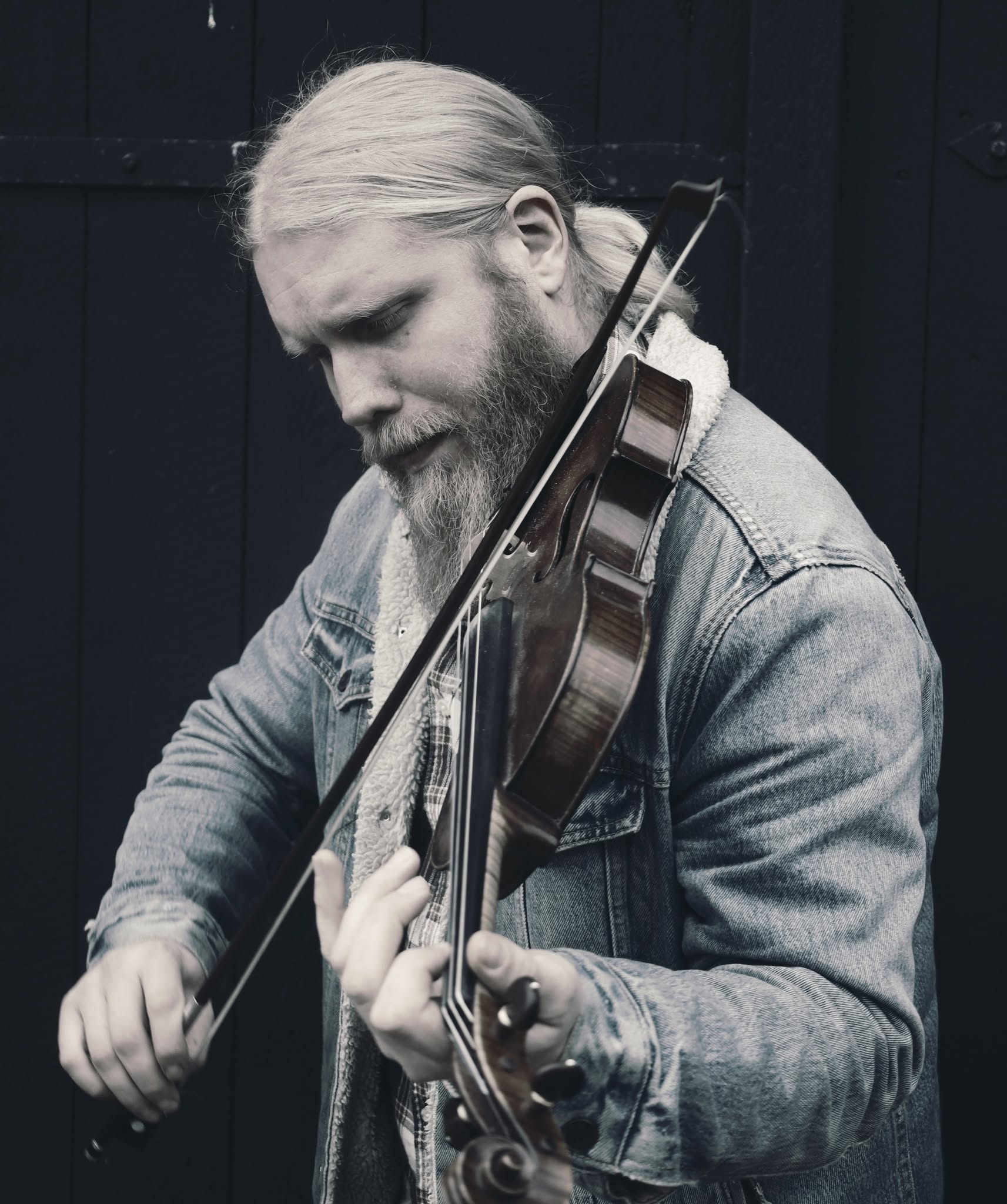 En man med långt hår och skägg spelar fiol iklädd en jeansjacka, helt uppslukad av sin musik.