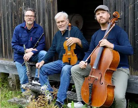 Tre män sitter utomhus vid en träbyggnad och håller i en klarinett, en fiol och en cello.
