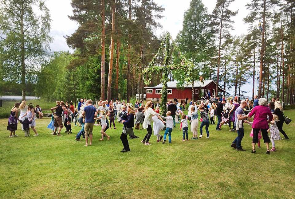 En grupp människor dansar utomhus runt en dekorerad majstång omgiven av träd och en röd stuga.