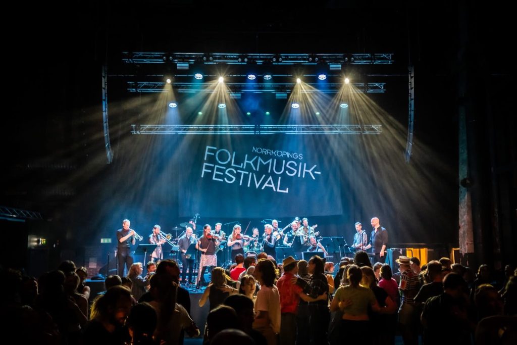 Ett band uppträder på scenen på Norrköpings folkmusikfestival medan folk dansar under färgglada lampor.