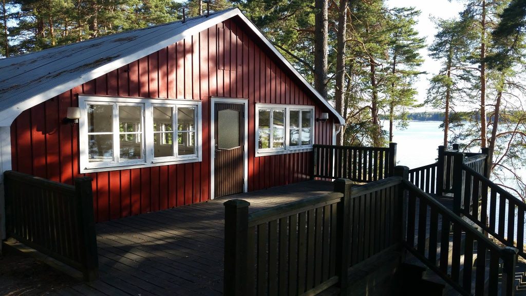 En röd trästuga med vitmålade fönster, omgiven av träd, med utsikt över en sjö i solskenet.